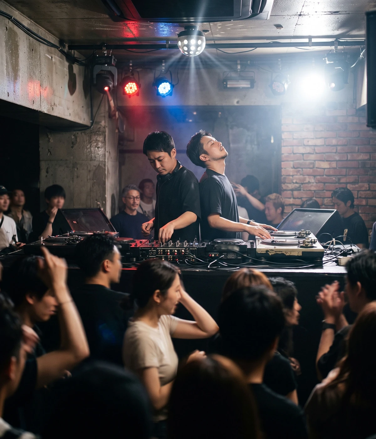 Two DJs performing back to back in a packed, dimly lit Tokyo club, colored stage lights cutting through haze, a dense crowd dancing in the foreground.